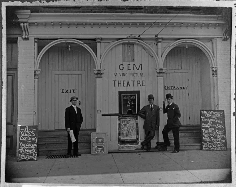 Three men in hats standing in front of the Gem Moving Picture Theatre. A chalkboard on each side of the door.