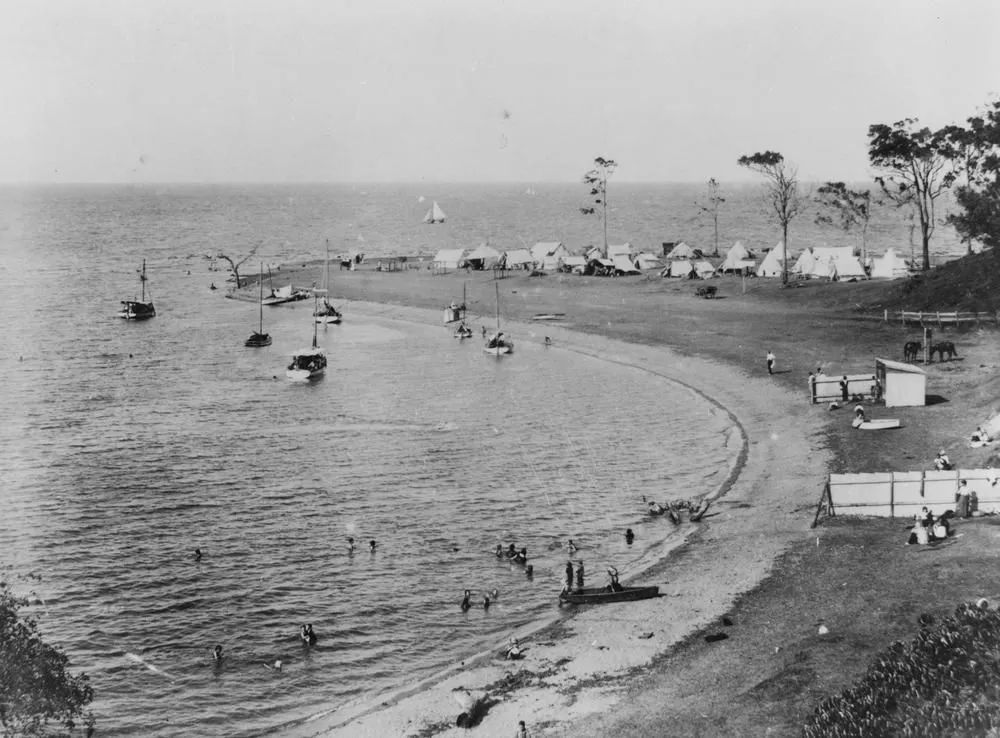 A black-and-white photograph of a curved beach. Boats float in the shallow water and white tents stand under gum trees.