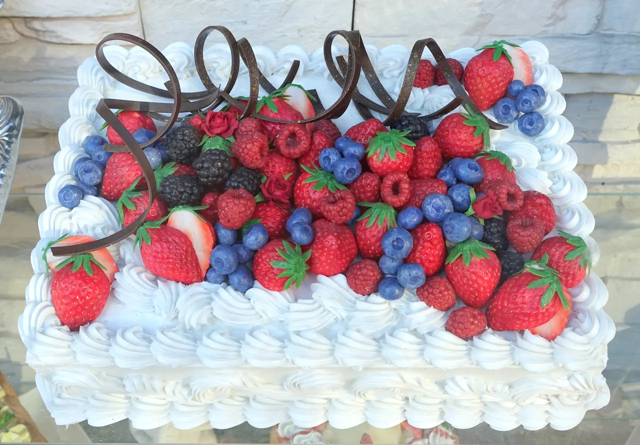A square white cake seen from above. Swirls of whipped cream cover the sides, and the top is heaped with strawberries, blueberries, raspberries and curls of dark chocolate.