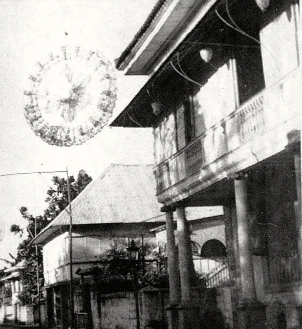 An old photograph of a street with a house on the right. A huge round star lantern with many points hangs over the road.